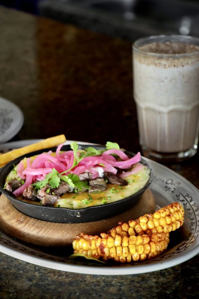 Platillo De Papa Cremosa Con Arrachera, Cebolla Morada Y Costillas De Elote, Acompañado De Una Bebida Fría En Un Restaurante.