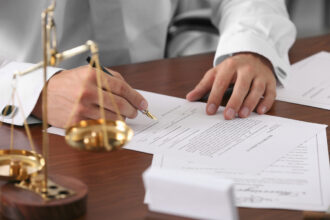 Primer Plano De Un Abogado O Notario Con Camisa Blanca Y Mancuernillas Firmando Documentos Legales Con Pluma Estilográfica Sobre Un Escritorio De Madera, Al Lado De Una Balanza De La Justicia Dorada.