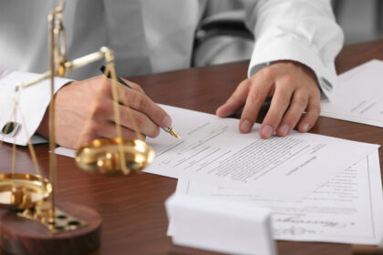 Primer Plano De Un Abogado O Notario Con Camisa Blanca Y Mancuernillas Firmando Documentos Legales Con Pluma Estilográfica Sobre Un Escritorio De Madera, Al Lado De Una Balanza De La Justicia Dorada.