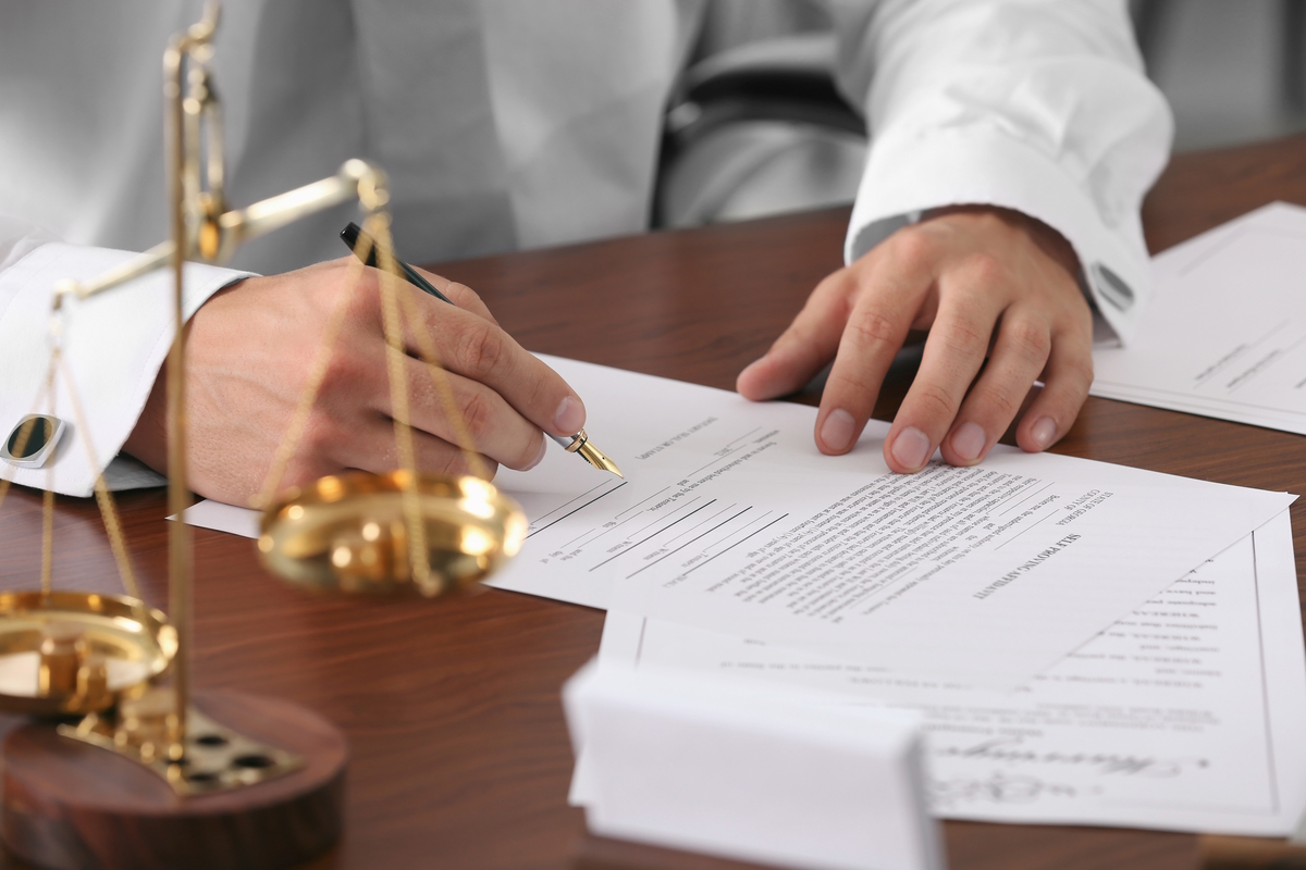 Primer Plano De Un Abogado O Notario Con Camisa Blanca Y Mancuernillas Firmando Documentos Legales Con Pluma Estilográfica Sobre Un Escritorio De Madera, Al Lado De Una Balanza De La Justicia Dorada.