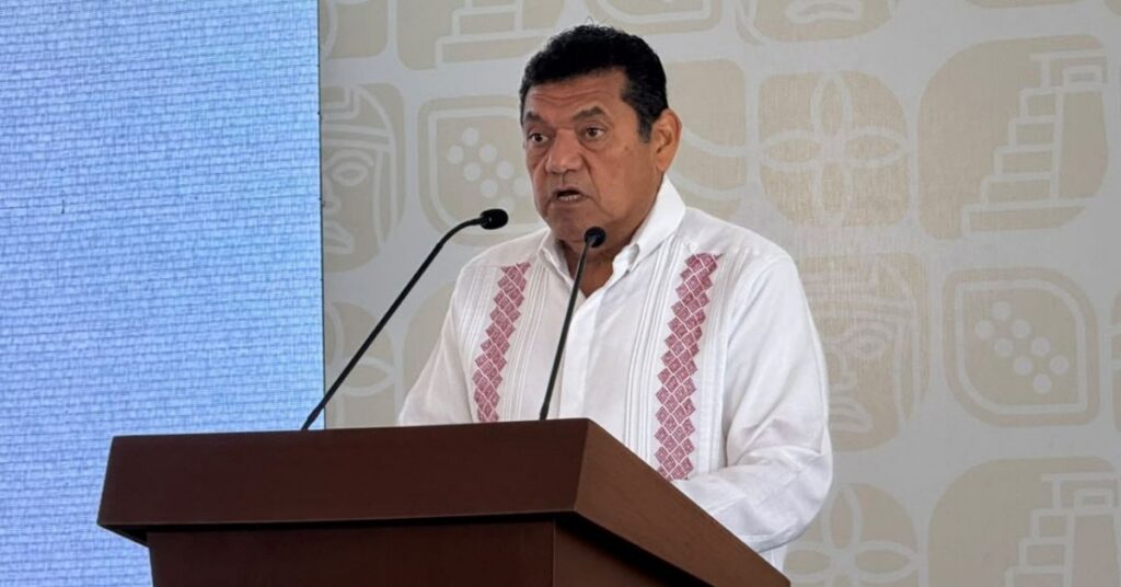 Respaldo Federal, Reforestación Masiva Y Multimillonaria Inversión Contra Inundaciones Marcan La Agenda En Tabasco Una Fotografía De Plano Medio Muestra A Un Hombre De Mediana Edad, Con Cabello Oscuro Y Corto, Hablando Detrás De Un Podio De Madera Oscura. Viste Una Guayabera Blanca Tradicional Con Bordados Detallados En Tonos Rosados Y Morados A Los Lados. Dos Micrófonos De Solapa Están Sujetos A La Solapa De Su Camisa. El Fondo Es Una Pared Con Un Patrón Sutil De Relieves Geométricos Y Glifos Mayas Estilizados. La Iluminación Es Brillante Y Uniforme.