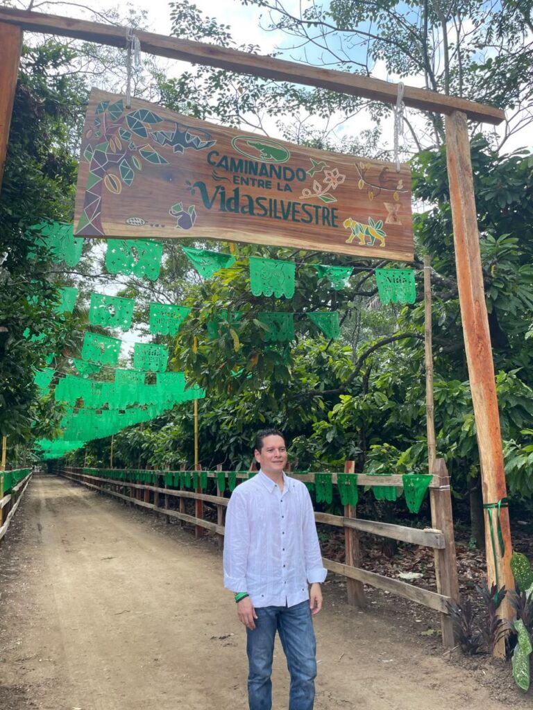 Cacao, Innovación Y Turismo: Inauguran La Experiencia “Caminando Entre La Vida Silvestre” En Cunduacán Para Impulsar El Desarrollo De Tabasco El Lic. Jesús Abraham Cano González De Pie Bajo Un Arco De Entrada De Madera Decorado Con Un Letrero Que Dice &Quot;Caminando Entre La Vida Silvestre&Quot;, Ubicado En Una Zona Natural Rodeada De Árboles Y Adornada Con Guirnaldas De Papel Picado Verde. Viste Una Guayabera Blanca Y Jeans Azules.