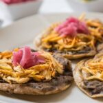 Primer Plano De Un Plato Blanco Con Tres Panuchos Tradicionales Servidos Con Base De Frijol, Carne Deshebrada Y Cebolla Morada Encurtida Encima.