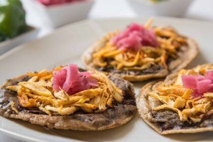 Primer Plano De Un Plato Blanco Con Tres Panuchos Tradicionales Servidos Con Base De Frijol, Carne Deshebrada Y Cebolla Morada Encurtida Encima.