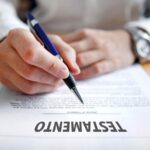 Primer Plano De Una Persona Sosteniendo Un Bolígrafo Azul A Punto De Firmar Un Documento Legal Que Lleva El Título &Quot;Testamento&Quot; En Letras Mayúsculas Sobre Un Escritorio De Madera.