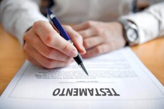 Primer Plano De Una Persona Sosteniendo Un Bolígrafo Azul A Punto De Firmar Un Documento Legal Que Lleva El Título &Quot;Testamento&Quot; En Letras Mayúsculas Sobre Un Escritorio De Madera.