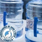 Garrafones Azules De Leonella Purificadora De Agua Alineados En Una Mesa Mientras Una Persona De Blusa Roja Se Acerca A Manipularlos