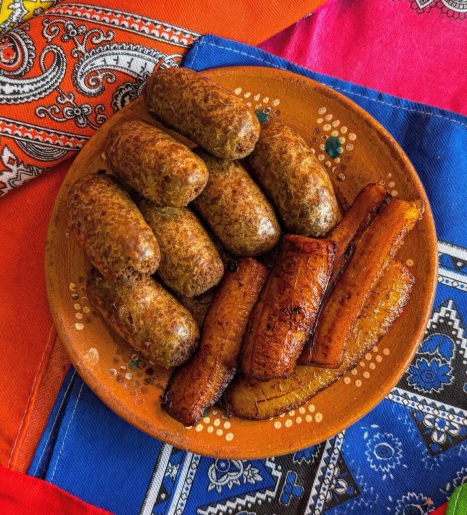 El Secreto Culinario De Jalpa: Tradición, Sabor E Identidad De La Butifarra Tabasqueña Plato De Barro Con Butifarras Tabasqueñas Asadas Y Plátanos Machos Fritos, Servido Sobre Un Mantel De Colores Vivos Con Patrones Tradicionales Mexicanos.
